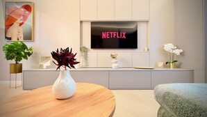 Una sala de estar moderna con un televisor de pantalla plana mostrando 'NETFLIX', un florero blanco con follaje oscuro, una mesa de centro de madera y un sofá verde.