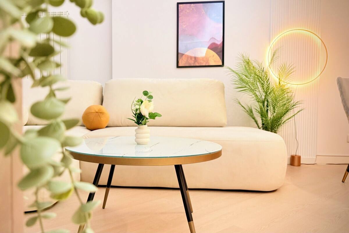Una sala de estar moderna con un sofá, una mesita de centro redonda de vidrio, un jarrón blanco con flores, un cojín amarillo y un cuadro en la pared.