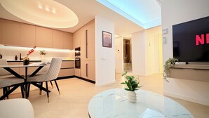 Una cocina moderna con electrodomésticos integrados, una zona de comedor con mesa y sillas blancas, y una sala de estar con televisor y planta.
