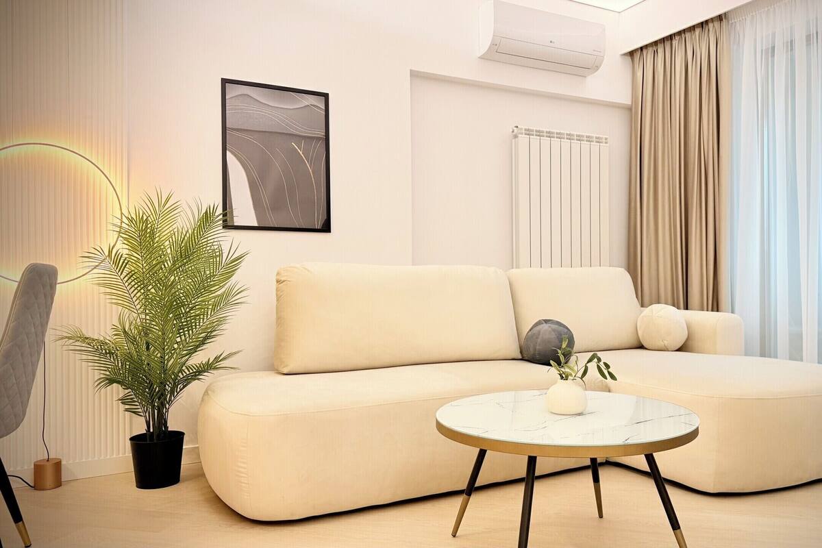 Una sala de estar moderna con un sofá color beige, una mesa redonda de centro, una planta en maceta y un radiador.