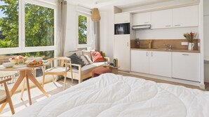 A modern bedroom with a large bed, a dining area with a table and chairs, a kitchenette, and a view of trees through large windows. - Le Barrel - studio for 3 - shared swimming pool (Biscarrosse)