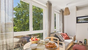 A modern living room with a sofa, dining table, and a balcony with a view of trees. - Le Barrel - studio for 3 - shared swimming pool (Biscarrosse)