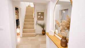 Ein Flur mit einer Holztreppe, einem Spiegel, einer Vase mit getrockneten Pflanzen und einer Tür mit einem Bulletinbrett.
