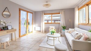 Uma sala de estar espaçosa, com sofá, mesa de centro redonda, um banquinho pequeno, escrivaninha com cadeira e uma janela grande com cortinas.
