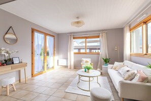 Ein geräumiges Wohnzimmer mit einer Couch, einem runden Couchtisch, einem kleinen Hocker, einem Schreibtisch mit Stuhl und einem großen Fenster mit Vorhängen.
