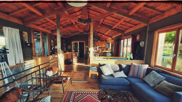 A spacious living area with a wooden ceiling, a blue sofa with cushions, a glass railing, and a view of the outdoors.