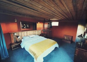 A bedroom with a wooden ceiling, a bed with a white and yellow blanket, a nightstand, a desk with a lamp, and a window.