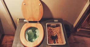 Un inodoro con asiento y tapa de madera, un recipiente de plástico con una cuchara, y piso de madera.