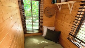 A small, cozy room with wooden walls, a bench, a bed, and a window with blinds.