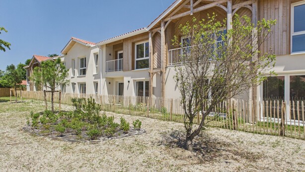 Un immeuble résidentiel à plusieurs étages, doté d’une façade en bois, de nombreux balcons et d’une cour clôturée avec une végétation clairsemée.