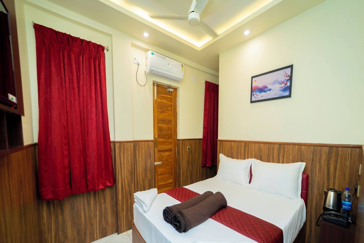 A hotel room with a bed, red curtains, a painting, a water bottle, and a coffee maker on a nightstand.