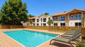 Une piscine avec une terrasse en bois, un grand arbre et un immeuble à plusieurs étages doté de balcons.
