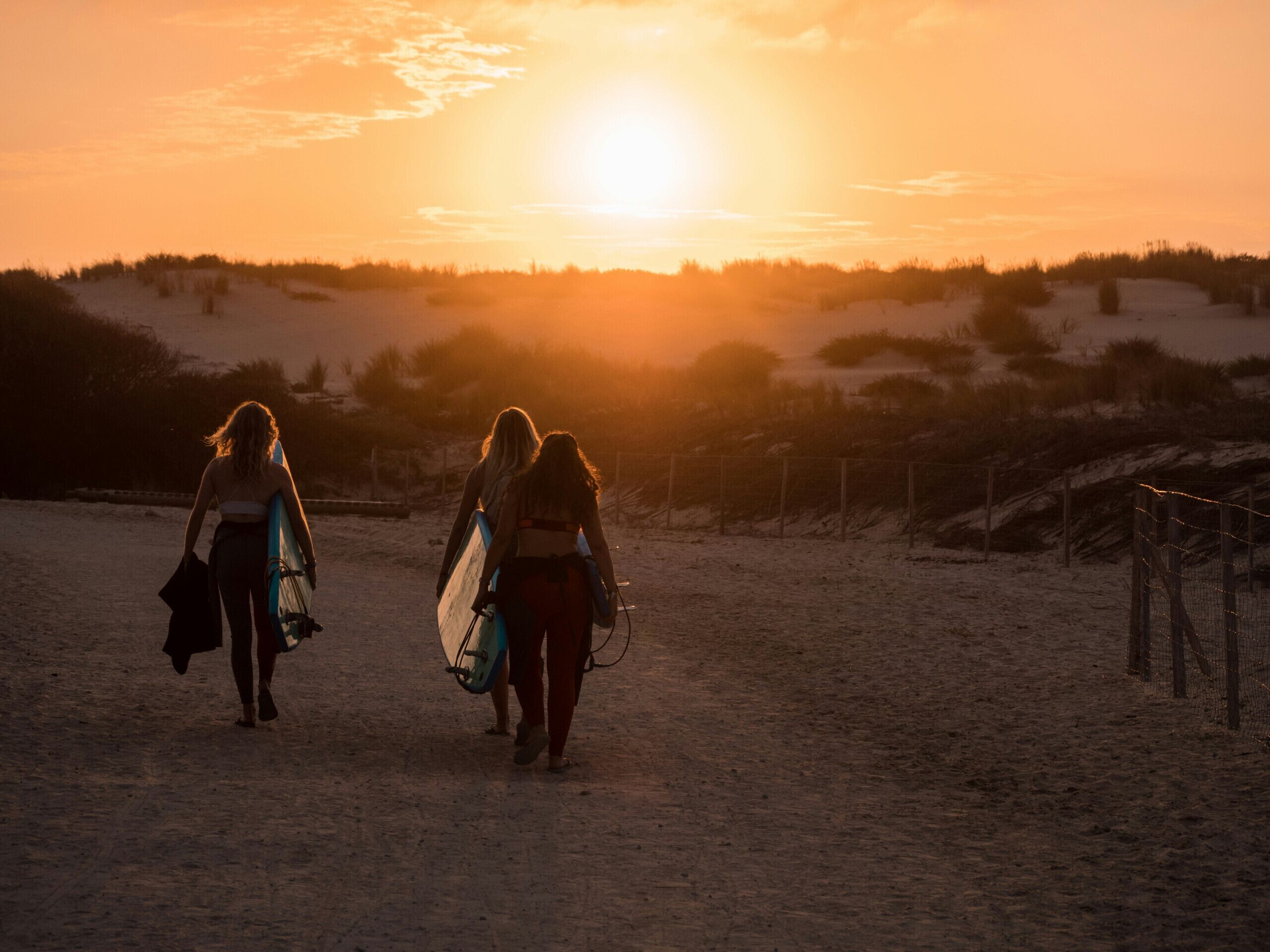 Deux personnes se promènent sur un sentier sablonneux, avec un coucher de soleil en arrière-plan.