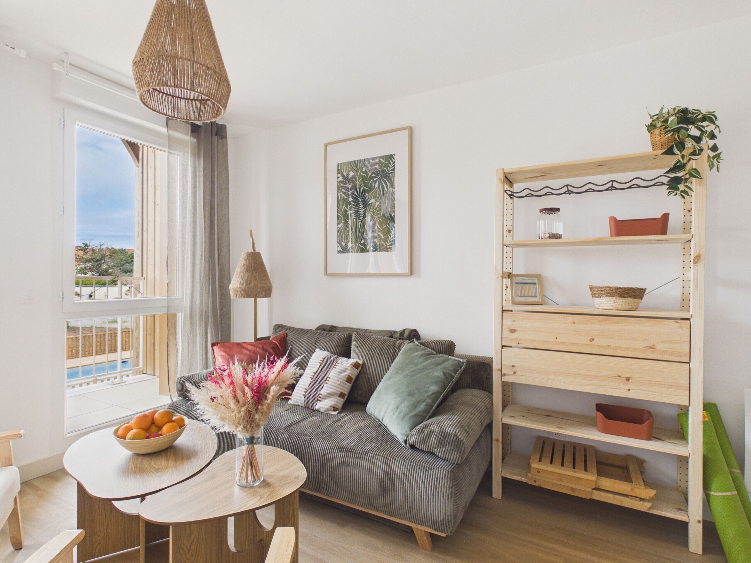A living room with a sofa, a table with a bowl of fruit, a hanging lamp, and a wooden shelf with decorative items.