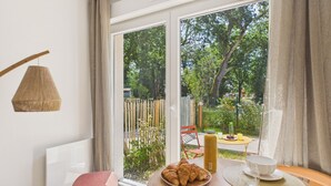 A modern room with a large sliding glass door leading to a garden, a small wooden table with a plate of croissants, a bottle, and two white cups. - The Board -apartment 4 ad 1 kid -shared swimming pool (Biscarrosse)