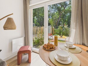 A modern room with a large sliding glass door leading to a garden, a small wooden table with breakfast items, a red stool, and a hanging light fixture. - The Board -apartment 4 ad 1 kid -shared swimming pool (Biscarrosse)