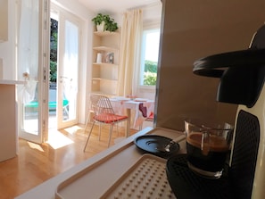A room with a dining table, a chair, a coffee machine, and a view of an outdoor area with a pool.