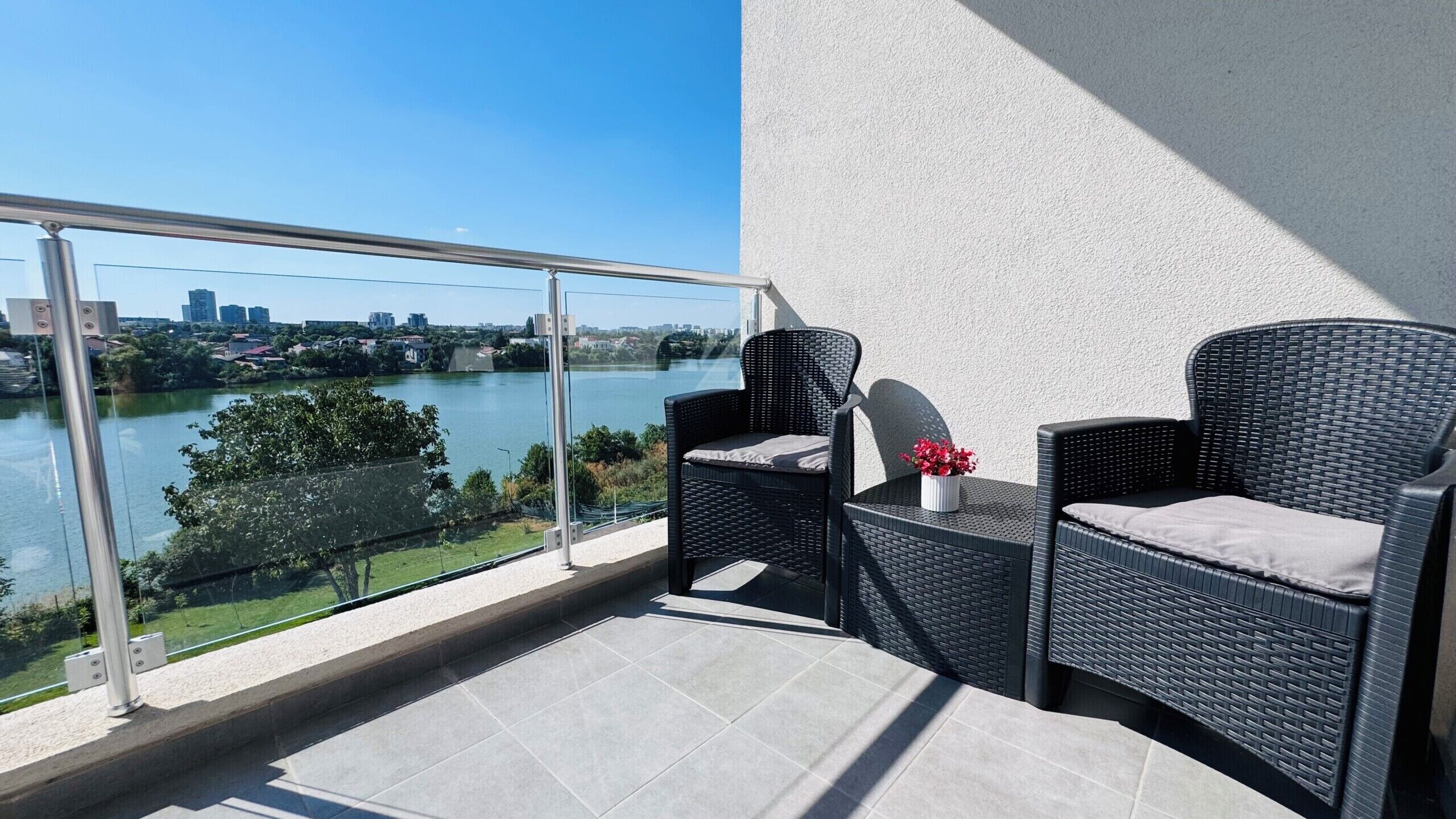 A balcony with two wicker chairs, a small table with a vase of flowers, and a view of a river and cityscape.