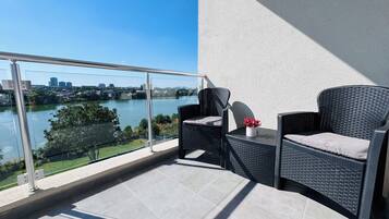 A balcony with two wicker chairs, a small table with a vase of flowers, and a view of a river and cityscape.
