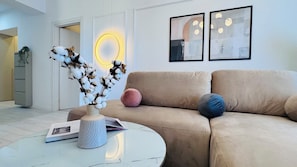 Un salon moderne avec un canapé beige, une table basse ronde et un vase décoré de branches de coton. Deux œuvres d’art encadrées ornent le mur, et un néon éclaire la pièce.