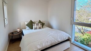 Een slaapkamer met een bed, nachtkastje, lamp en een raam met uitzicht op groen.