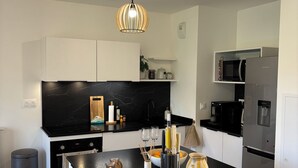 Een moderne keuken met een centraal eiland, zwarte aanstootbladen, witte kastjes, een zwarte kookplaat, een magnetron, een koelkast en een hanglamp.