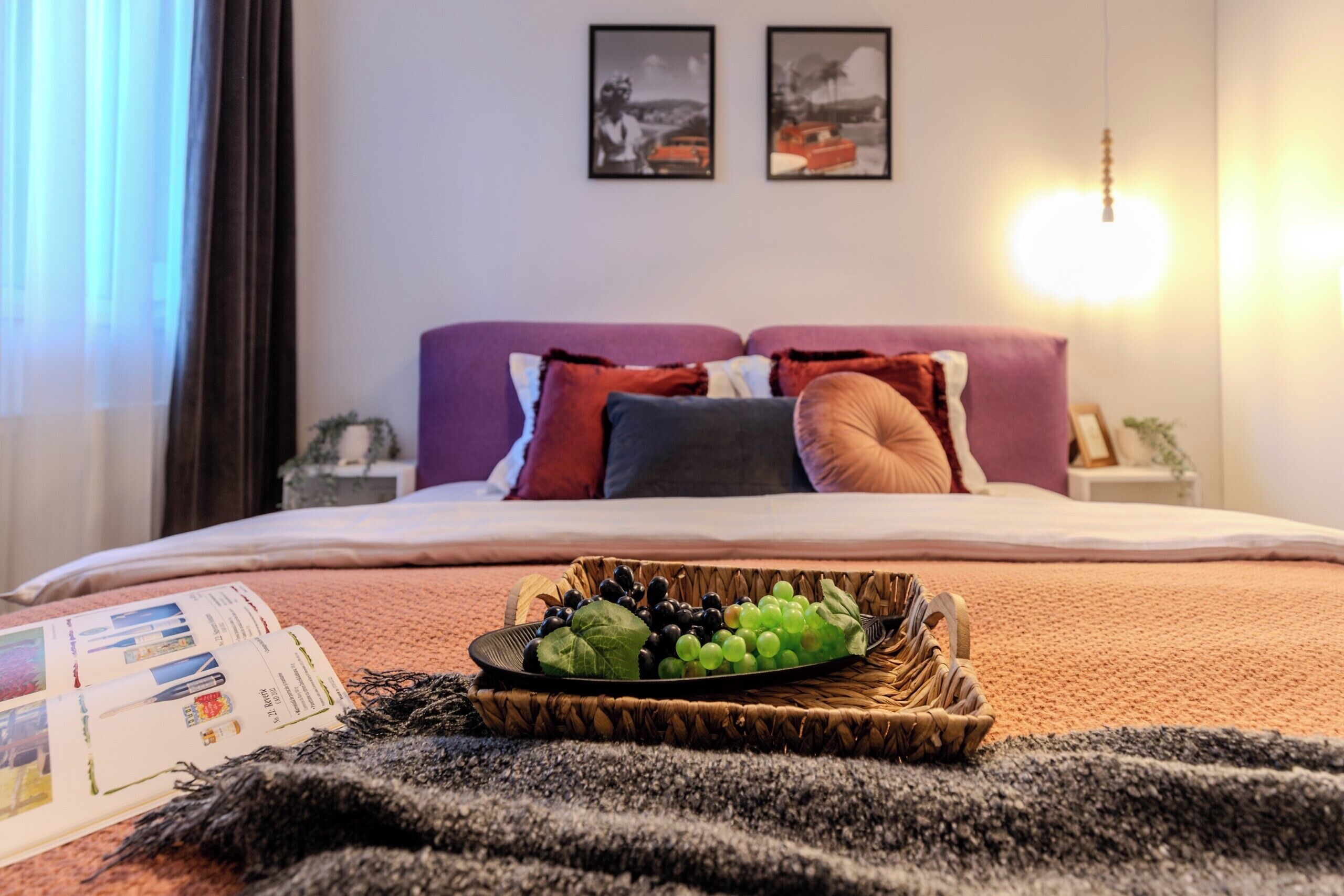 Una cama bien hecha con una cesta de fruta, una revista y una lámpara sobre una mesita de noche.