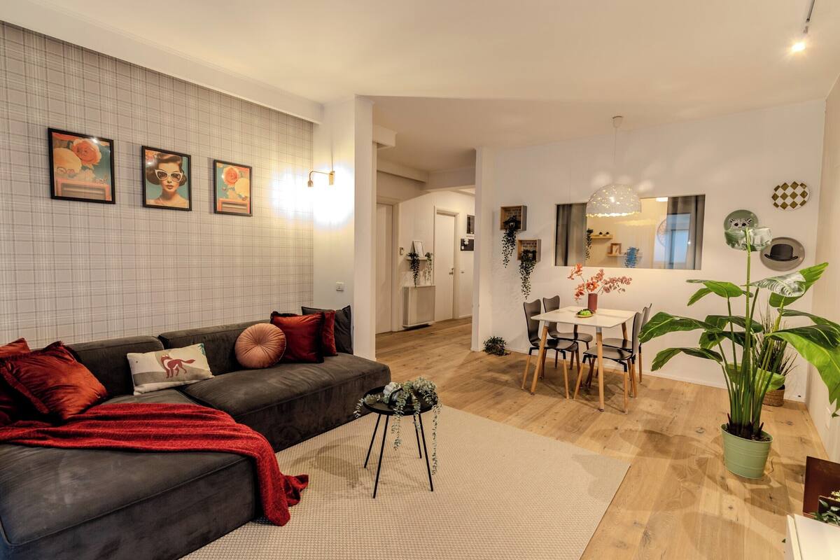 Una sala moderna con un sofá oscuro, cojines rojos, una mesita auxiliar y una zona de comedor con mesa y sillas.