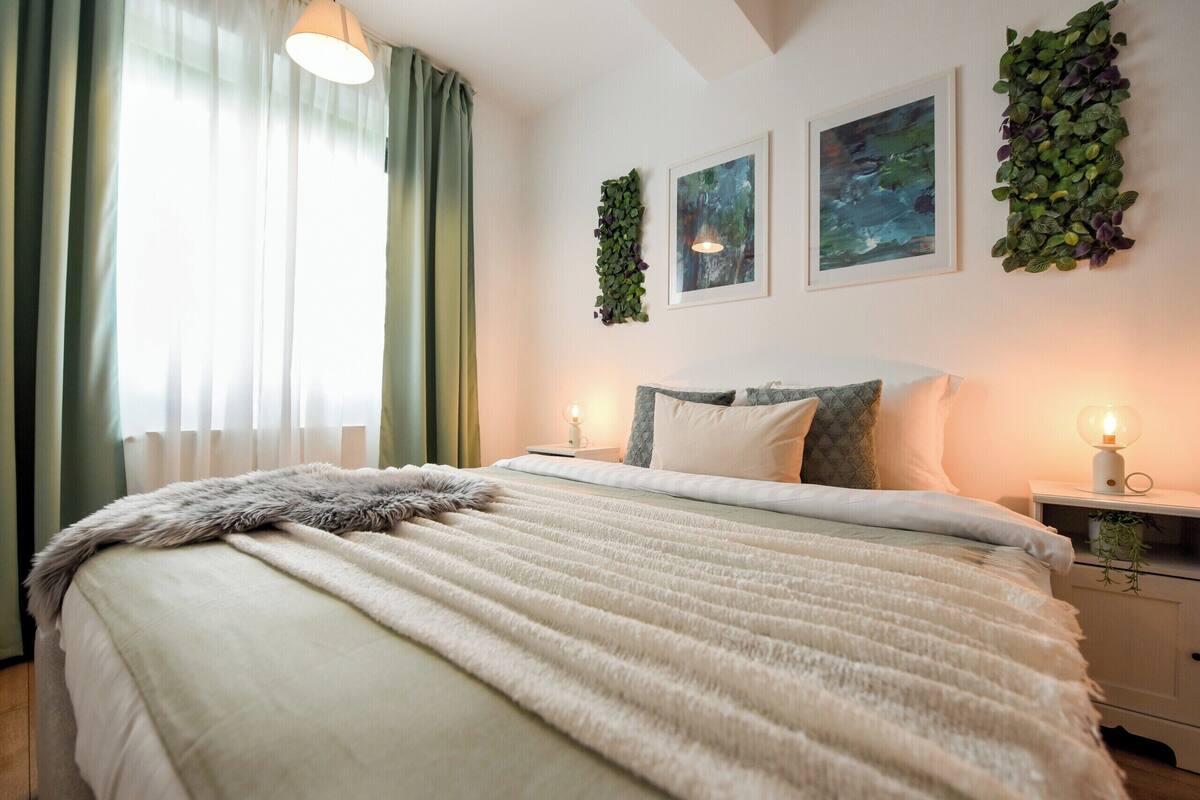 Un dormitorio con cama, lámparas de noche, ventana con cortinas y cuadros en la pared.