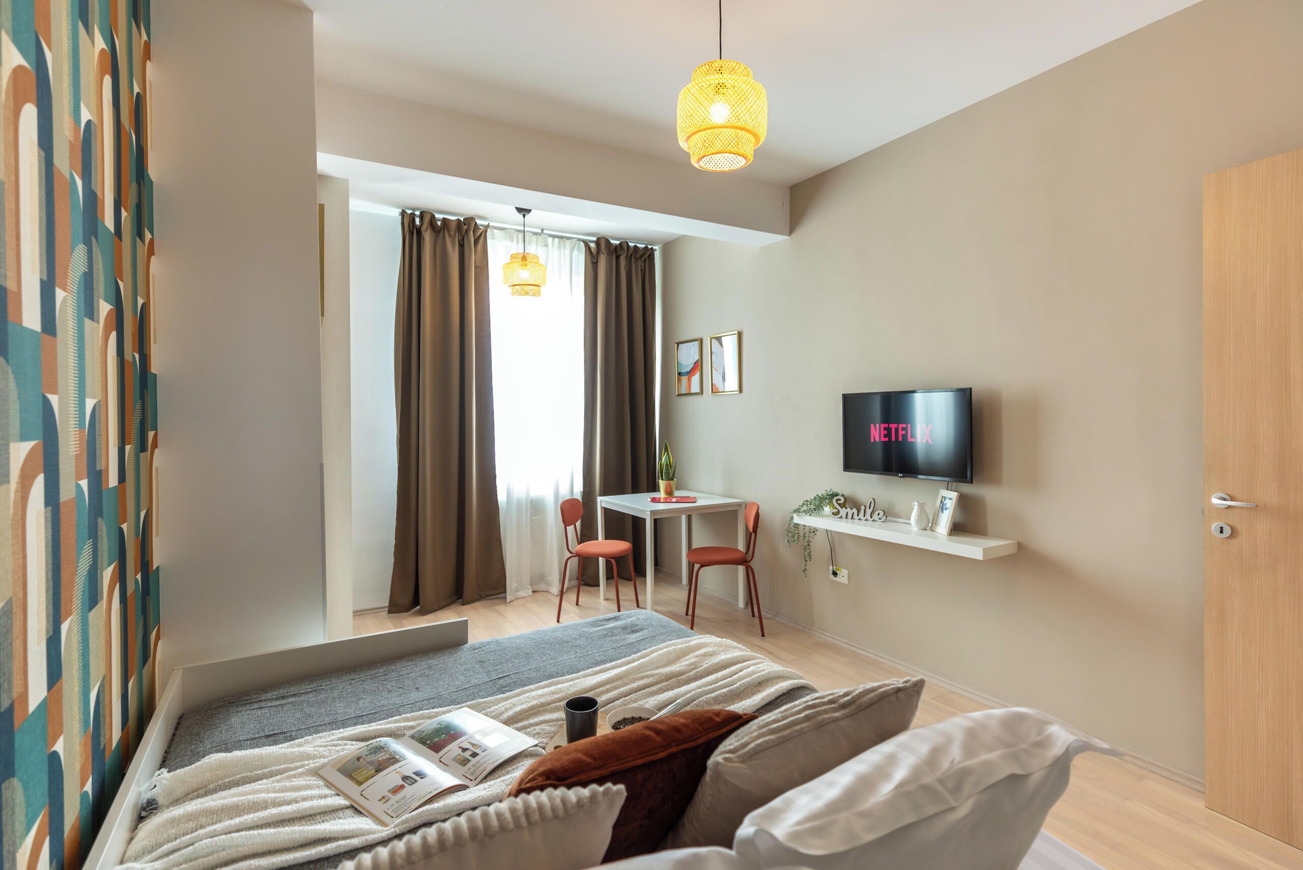 Un dormitorio moderno con cama, televisor, escritorio y sillas, y ventana con cortinas.