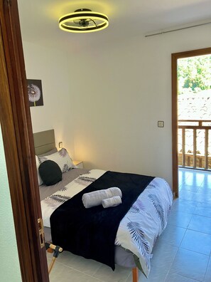 Ein Schlafzimmer mit Bett, Nachttisch, Deckenventilator und Fenster mit Blick auf Grünflächen.