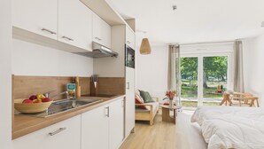 Eine moderne Küche mit weißen Schränken, einer Holzausziehtafel und einer Obstschüssel. Es gibt einen Essbereich mit Tisch und Stühlen sowie ein Bett mit weißer Bettdecke.