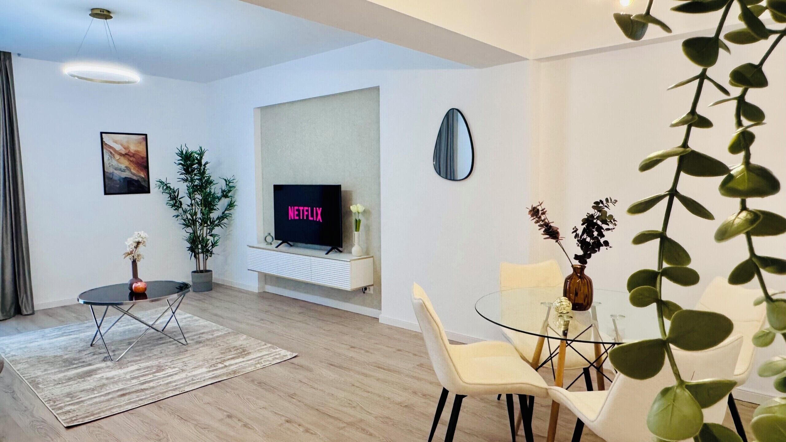 Una sala de estar moderna con un televisor de pantalla plana, una mesa de comedor con sillas, una pequeña mesa redonda con un florero y una planta en maceta.