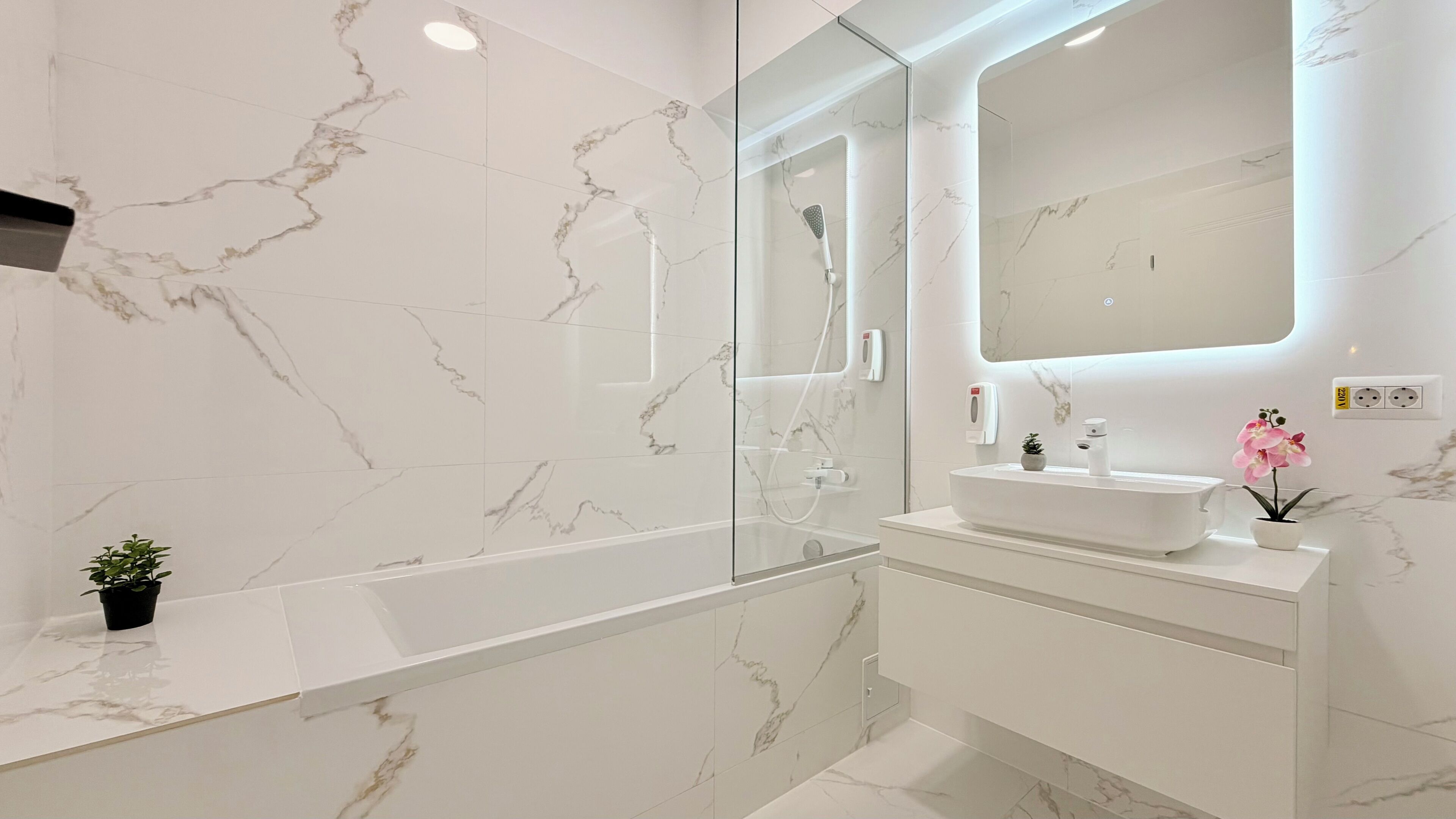 Un baño moderno con suelo de mármol, una bañera independiente, un espejo grande y un lavamanos blanco con una pequeña planta en maceta.
