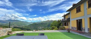 A villa with a view of mountains, a patio with artificial grass, and a yellow building with a balcony.