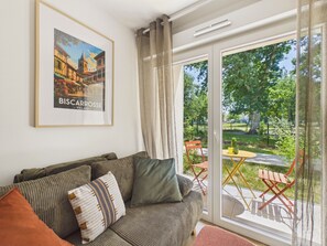 Un salon avec un canapé, un tableau encadré représentant Bischkowerse et une vue sur un jardin, visible à travers une porte vitrée.