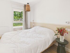 A bedroom with a bed, a window, a hanging light, and a vase with flowers. - La Planche Malibu - apartment for 4- shared pool (Biscarrosse)