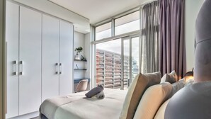 Ein modernes Schlafzimmer mit einem großen Bett, einem Kleiderschrank, einem Schreibtisch und Blick auf die Skyline durch große Fenster.