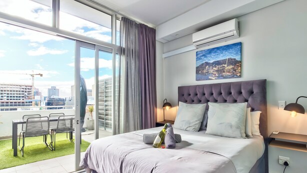 Ein Schlafzimmer mit einem großen Bett, Blick auf die Stadt durch eine Schiebetür, einem Balkon mit Sitzgelegenheiten und einem Stadtbild an der Wand.