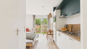 A modern kitchen with a large island, a dining area with a table and chairs, and a bedroom with a bed and a view of the garden. - Le Funboard - studio for 3 - shared pool (Biscarrosse)