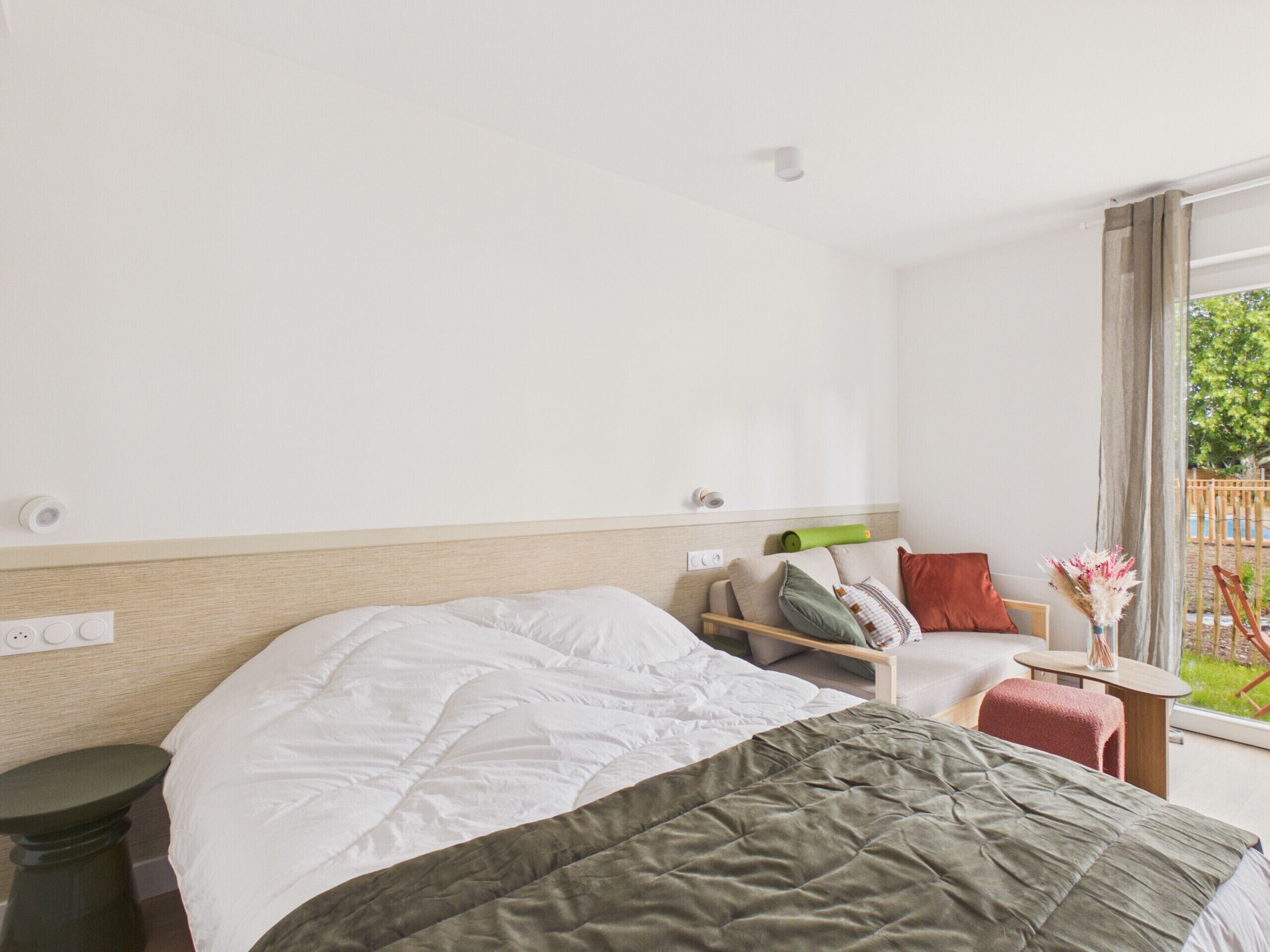 A hotel room with a bed, a small table, a chair, and a window with a view of greenery.