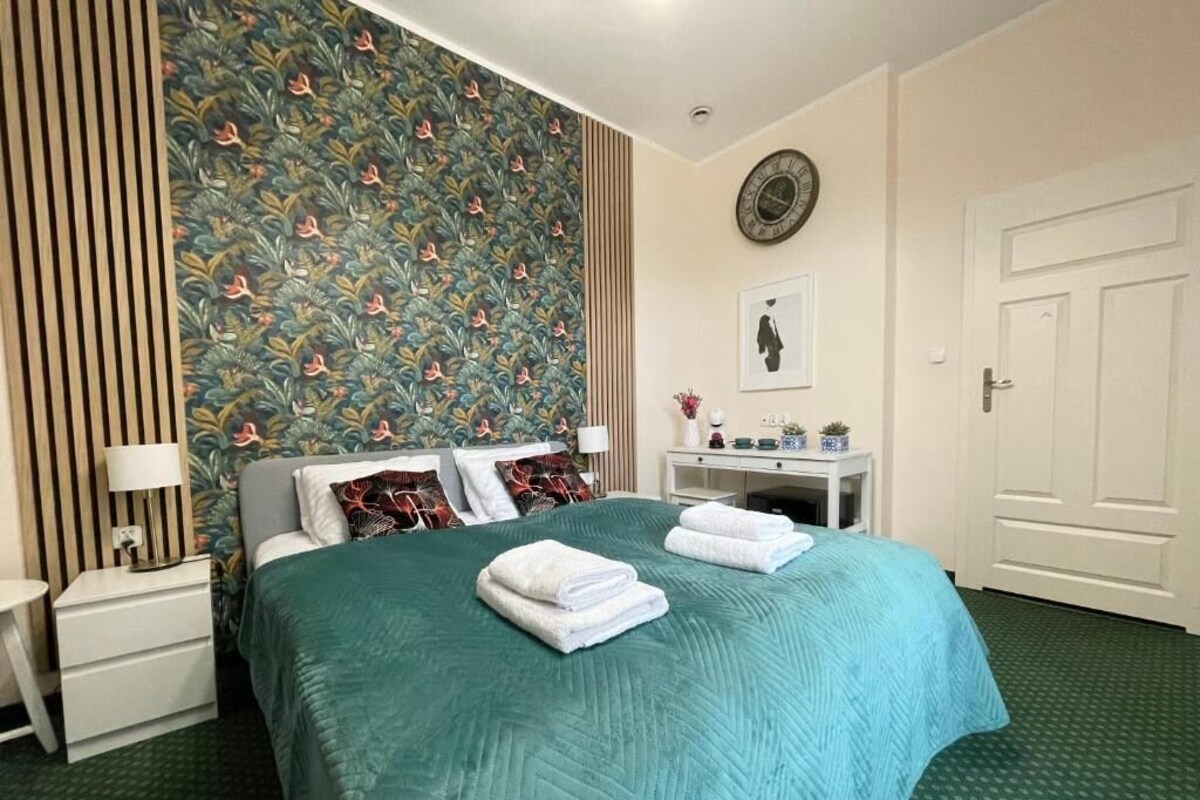 Quarto de hotel bem arrumado com cama, mesa de cabeceira, relógio de parede e porta.