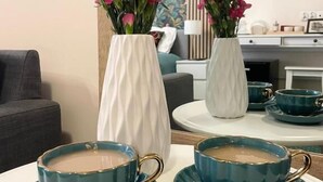 Een eetgedeelte met twee witte vazen vol roze bloemen, een tafel met vier turquoise kopjes en schoteltjes, en een grijze bank op de achtergrond.