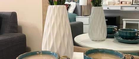 A dining area with two white vases of pink flowers, a table with four teal cups and saucers, and a grey sofa in the background.