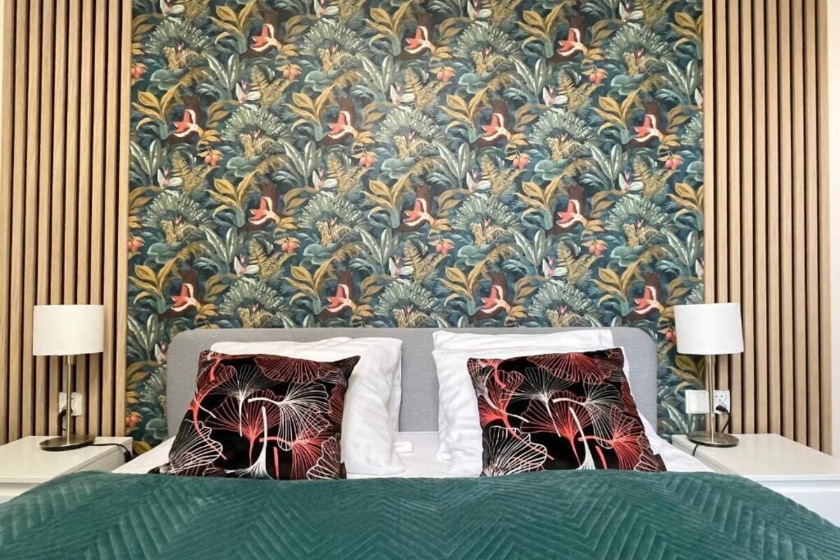Quarto de hotel com cama, duas mesas de cabeceira, um criado-mudo, parede com padrão floral e colcha verde.