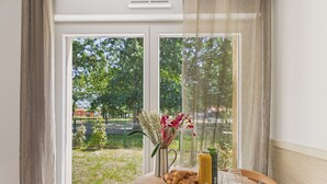 A room with a table set for breakfast, a window with a view of greenery and a building, and a curtain partially drawn. - Le Nose Rider - studio for 3 - shared pool (Biscarrosse)