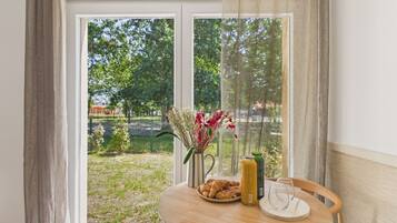 A room with a table set for breakfast, a window with a view of greenery and a building, and a curtain partially drawn.