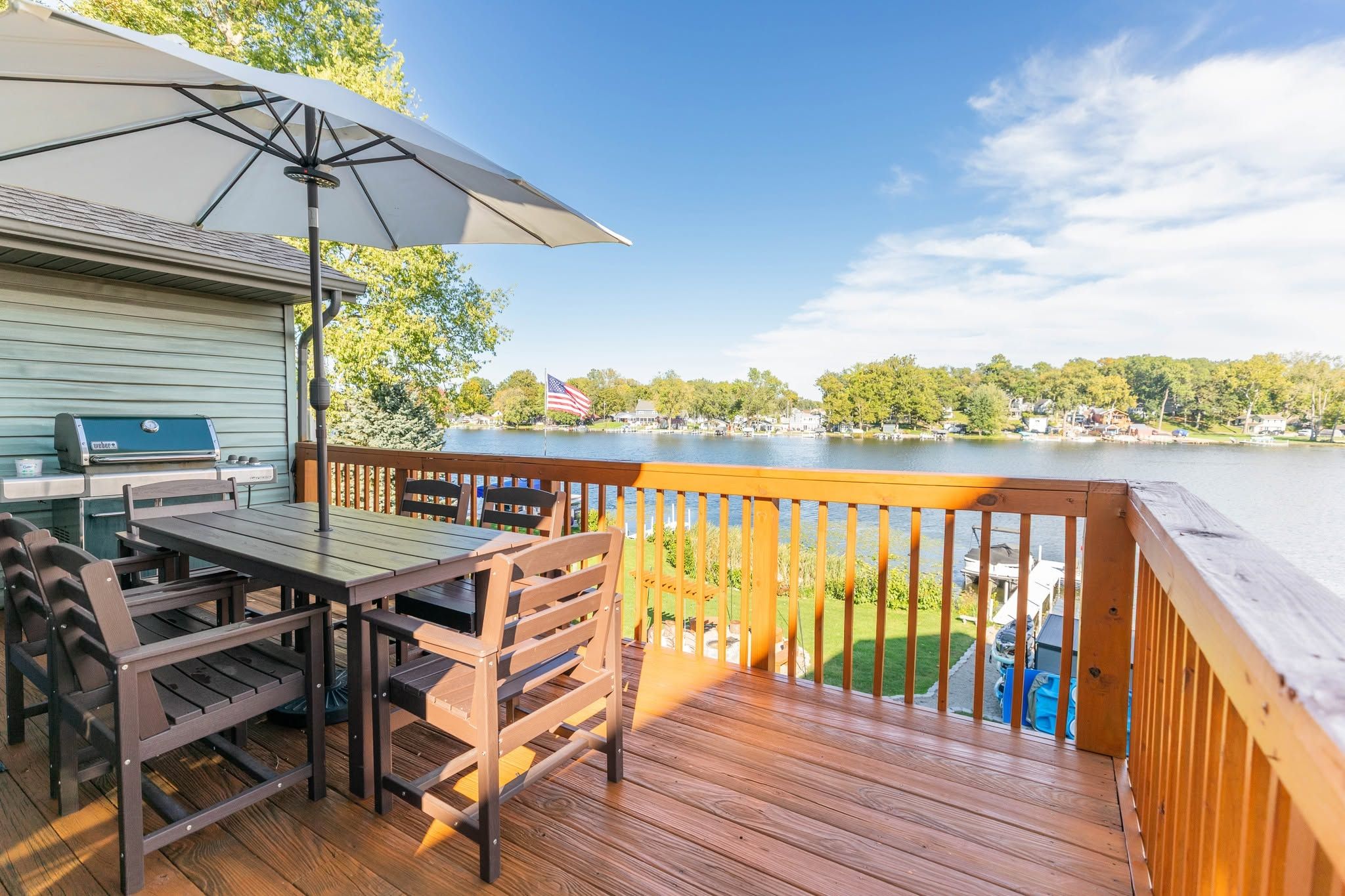 Een houten terras met een barbecue en tafel, met uitzicht op een meer, huizen en bomen.