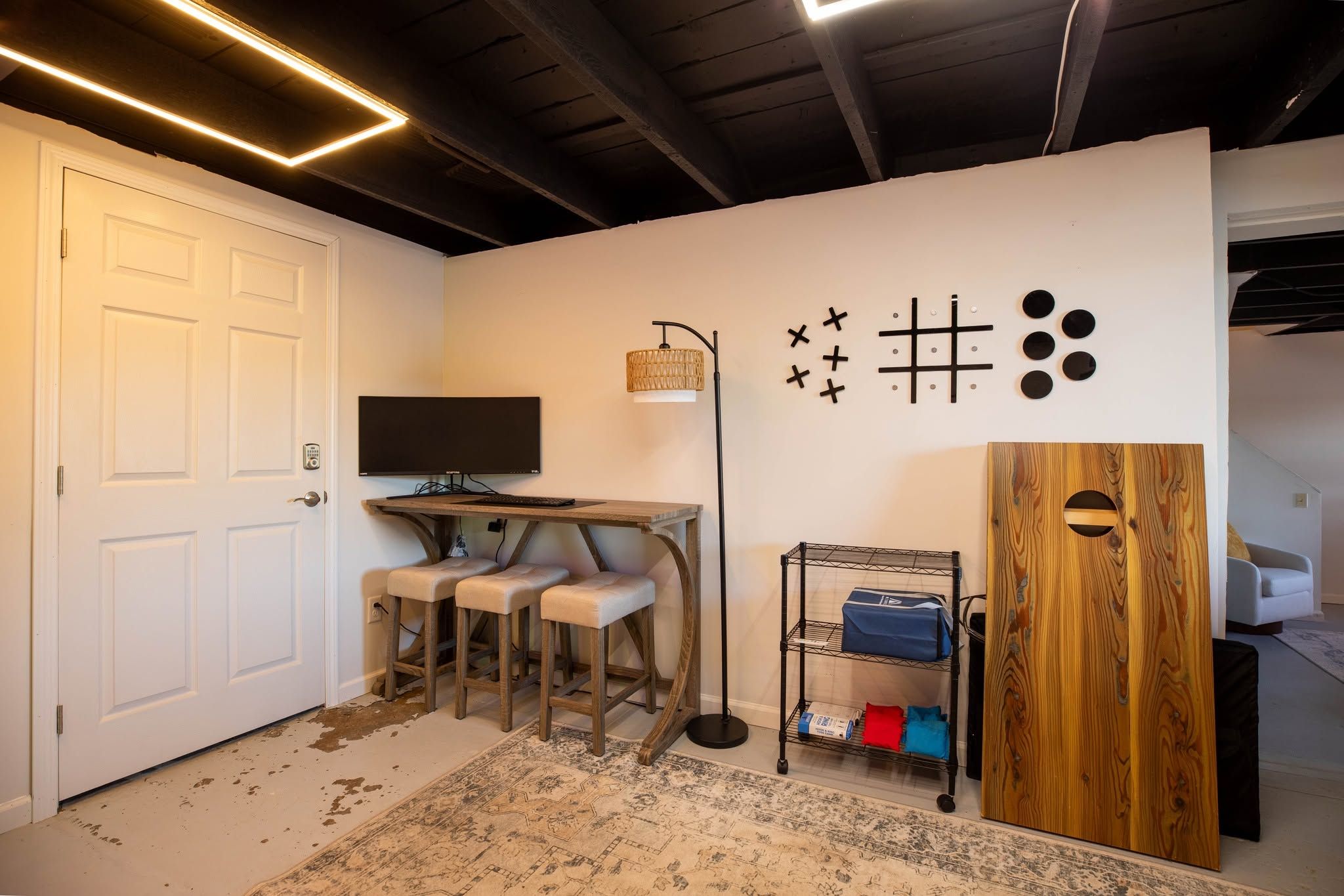 A room with a white door, a desk with a computer, two chairs, a floor lamp, a wall with abstract art, and a wooden cabinet.