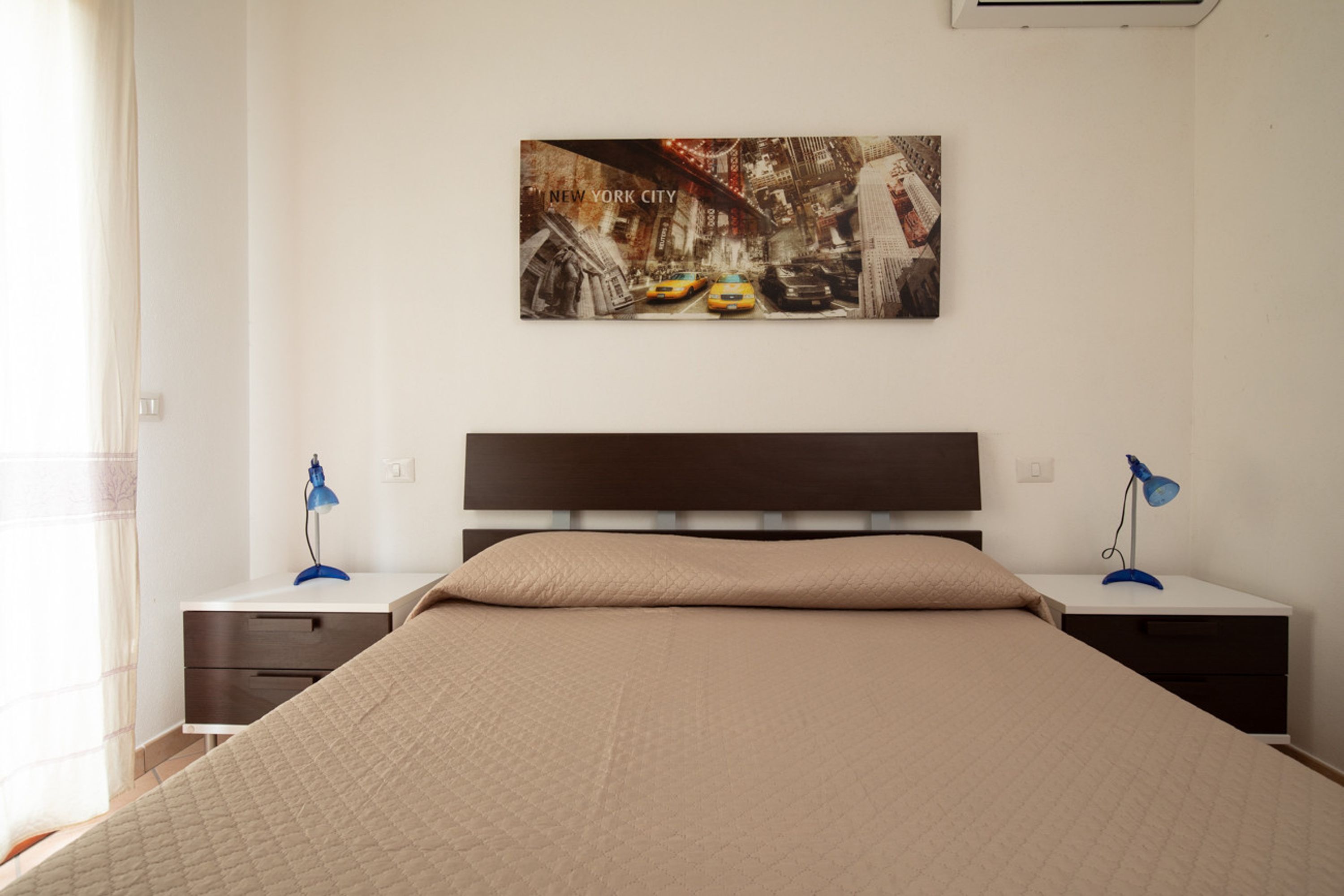 A bedroom with a bed, a nightstand with a lamp, a wall-mounted picture, and a window with curtains.